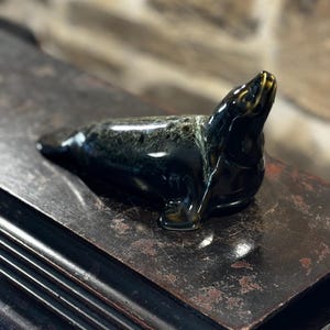 Puede incluir: Una figurita de foca de cerámica negra y verde con un acabado brillante. La foca está tumbada sobre su vientre con la cabeza levantada y mirando hacia la derecha. La figurita está sentada sobre una superficie de madera oscura.