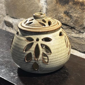 May include: A ceramic candle holder with a lid. The holder is a light beige colour with brown accents and leaf-shaped cutouts. The lid has several small, oval-shaped cutouts. The candle holder is round and sits on a dark surface.