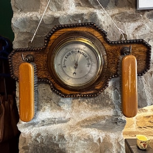 May include: An antique wooden barometer with a circular gauge and two wooden handles. The gauge has a glass cover and displays weather conditions. The wooden frame is adorned with decorative elements. The barometer is mounted on a stone wall.