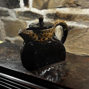 Pode incluir: Uma chaleira de cerâmica preta com tampa e alça. A borda superior e a alça têm um padrão marrom e bege salpicado. A chaleira tem um design decorativo em relevo na frente.