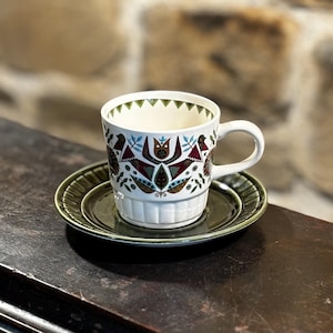 Könnte beinhalten: Eine weiße Kaffeetasse aus Keramik mit einem geometrischen Muster in Grün und Braun. Die Tasse hat einen Henkel und steht auf einer passenden grünen Untertasse.