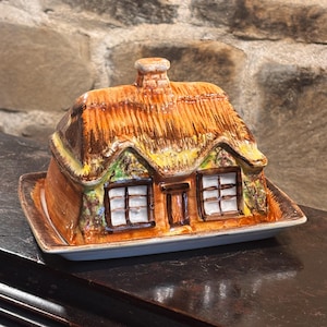Peut inclure: Un beurrier en céramique en forme de cottage avec un toit de chaume, une cheminée centrale et deux fenêtres. L'extérieur présente des détails peints en marron, jaune et vert sur une base rectangulaire, conçu pour contenir du beurre.