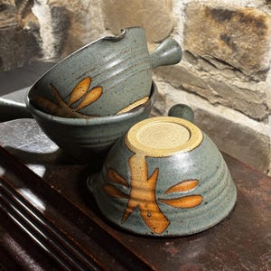 Puede incluir: Tres cuencos de sopa de cerámica hechos a mano apilados. Los cuencos son de color verde azulado apagado con un diseño floral marrón. Cada cuenco tiene un asa. Los cuencos están sobre una superficie de madera oscura.