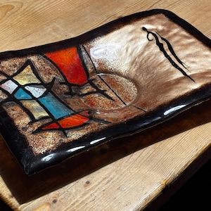 May include: A rectangular ceramic dish with a black border and a brown interior. The dish has an abstract design with red, blue, yellow, and black shapes. A black silhouette of a figure is on the right side.