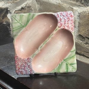 May include: A square, ceramic serving dish with two elongated, light pink compartments. The dish is decorated with green leaves and clusters of pink and white flowers. The dish is displayed on a black surface.