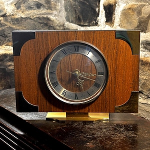 Puede incluir: Un reloj rectangular de madera con una esfera redonda. La esfera tiene números romanos y agujas doradas. El reloj tiene detalles dorados en las esquinas y una base dorada. Ancho aproximado: 20 cm.