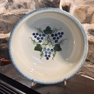May include: A round ceramic bowl with a cream-colored interior featuring a central hand-painted design of three clusters of blue grapes and green leaves. The rim of the bowl has a speckled blue and white border, providing a textured appearance.