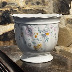 May include: White ceramic planter with a scalloped rim and a pedestal base. The planter is decorated with a floral pattern in shades of pink, yellow, and blue. The rim has a gold trim.