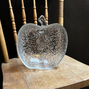 May include: A clear glass apple-shaped serving dish with a textured surface. The dish is sitting on a wooden chair.