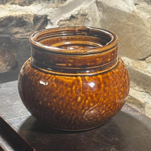 Puede incluir: Un pequeño tarro redondo de cerámica marrón con una superficie texturizada. El tarro tiene una abertura ancha y un borde ligeramente elevado. El exterior presenta un patrón moteado y un acabado brillante. El tarro es probablemente para almacenamiento o fines decorativos.