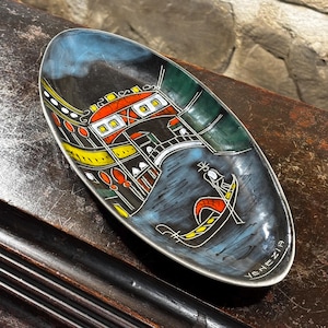 May include: An oval-shaped ceramic dish with a black background and a colorful illustration of a gondola in Venice, Italy. The dish is decorated with red, yellow, green, and blue paint. The text "VENEZIA" is written in white on the bottom right corner of the dish.