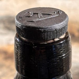 Puede incluir: Un cuello de botella de vidrio marrón oscuro rematado con una tapa circular negra que presenta un borde estriado texturizado y el número 27 en relieve en la superficie superior. La botella tiene un acabado liso y reflectante y un corcho visible.