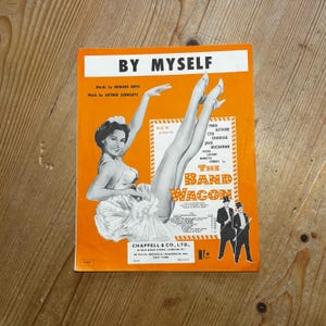 Könnte beinhalten: Ein Vintage-Notenblattcover für das Lied "By Myself" aus dem Musical "The Band Wagon". Das Cover zeigt ein Schwarzweißfoto einer Frau in einem weißen Tutu, mit dem Titel des Liedes und des Musicals in Schwarzweiß auf orangefarbenem Hintergrund.