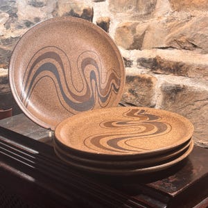 May include: A collection of brown ceramic plates with a speckled finish. Each plate has a wavy, abstract design in shades of brown and grey. The plates are stacked and displayed on a dark wooden surface, with a stone wall in the background.