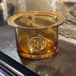 May include: An amber glass container with a wide rim and a clear, circular floral design on the side. The cylindrical container is made of amber glass, and the design is clear. The glass has a warm, inviting glow.