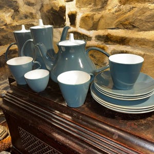 Könnte beinhalten: Ein teal-farbenes Teeservice mit weißen Innenbechern und Deckeln. Das Set umfasst eine Teekanne, einen Sahnekännchen, eine Zuckerdose, Tassen und Untertassen. Das Teeservice steht auf einer dunklen Holzoberfläche.