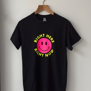 May include: Black t-shirt with a pink smiley face and the text "RIGHT HERE RIGHT NOW" in yellow.