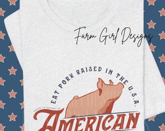 May include: A light gray t-shirt featuring a graphic of a pig and the text "EAT PORK RAISED IN THE U.S.A." in a circular design. Below, the shirt reads "AMERICAN TRADITION". The shirt also has the text "Farm Girl Designs".