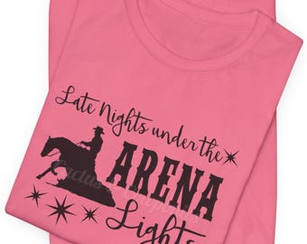 May include: Pink t-shirt with a black silhouette of a horse and rider, and the text "Late Nights under the ARENA Lights". The text "Cactus & Cowponies" is in the bottom right corner.