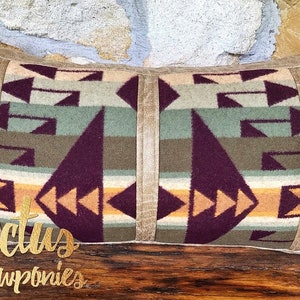 May include: A rectangular pillow with a southwestern pattern in shades of brown, green, purple, and orange. The pillow has a suede border and is sitting on a wooden surface. The text "Cactus & Cowponies" is visible in the bottom left corner of the image.