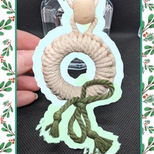 Op de afbeelding: Een witte macramé krans met een groene touw strik. De krans hangt aan een houten kraal.