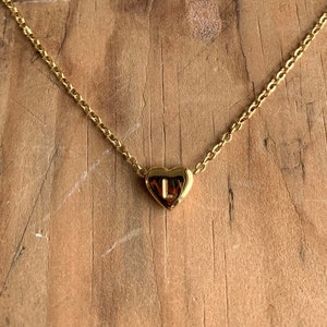 May include: A gold chain necklace with a small heart pendant featuring the letter "L" inside. The necklace is on a wooden surface.