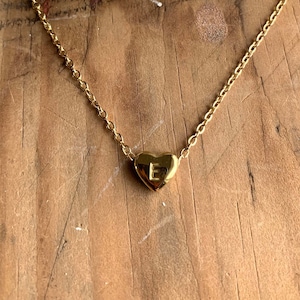 May include: A gold-colored necklace with a heart-shaped pendant. The pendant has the letter "E" engraved on it. The necklace is displayed on a wooden surface with white coral-like objects in the background.