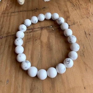 May include: White and gray marbled stone bead bracelet. The round beads are strung together to form a circular bracelet. The bracelet is displayed on a wooden surface with several white starfish.