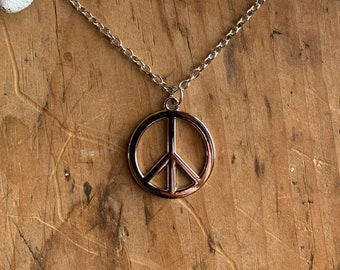 May include: A silver peace sign pendant necklace on a silver chain. The necklace is displayed on a wooden surface.
