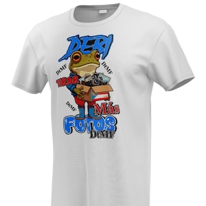 Può includere: T-shirt bianca con un cartone animato di una rana che tiene una scatola con macchine fotografiche e la scritta "DERI TIRAR MÁS FOTOS DIMF" in blu e rosso. La rana indossa stivali blu e una giacca blu.
