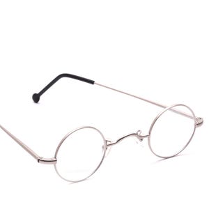 Könnte beinhalten: Runde Brille mit einem dünnen, silberfarbenen Metallrahmen und einem hohen Steg. Die Bügel sind schmal und gerade und enden in schwarzen, abgerundeten Kunststoffenden. Die klaren Gläser sind kreisförmig und das Design ist schlicht und leicht.