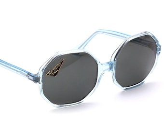 May include: Light blue, octagonal-shaped sunglasses with dark gray lenses. The frames are transparent, and a small tag reads "HEAT TREATED LENSES". A stylish accessory for eye protection.