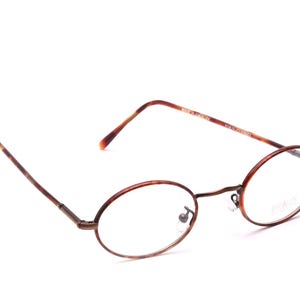 Può includere: Occhiali di forma ovale con un motivo marrone tartarugato sulle aste e sulla montatura. Le lenti sono trasparenti e la montatura è in metallo marrone scuro. Gli occhiali sono su uno sfondo bianco.
