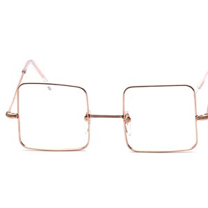 Peut inclure: Lunettes à monture carrée en métal or rose. Les lunettes ont un design minimaliste avec une monture fine et des verres transparents. Les branches et le pont de nez sont également en or rose. Les lunettes sont posées sur un fond blanc.