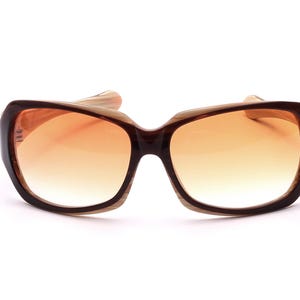 Könnte beinhalten: Sonnenbrille mit braunem Rahmen und orangefarbenen Gläsern mit Farbverlauf. Die Sonnenbrille hat eine breite, rechteckige Form mit leicht gebogener Oberseite. Die Bügel sind hellbraun. Die Gläser gehen von dunklerem Orange oben zu hellerem Orange unten über.