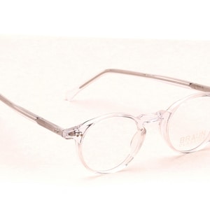 Könnte beinhalten: Transparente Brille mit runden Gläsern und silberfarbenen Bügeln. Die Brille hat ein minimalistisches Design und ist vor einem weißen Hintergrund platziert. Der Markenname "BRAUN OPTIK" ist auf dem Glas sichtbar.