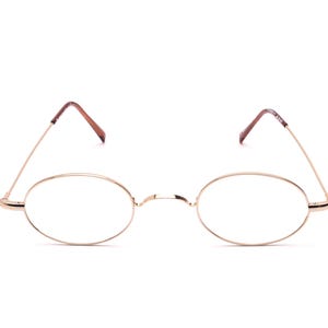 Peut inclure: Lunettes ovales dorées avec des verres transparents. La monture est fine et délicate, avec un pont reliant les deux verres. Les branches sont marron. Les lunettes sont posées sur un fond blanc.