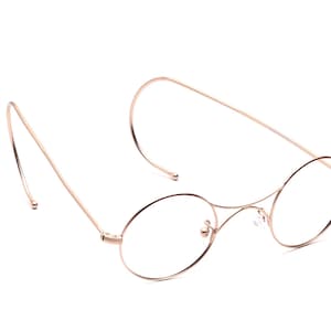 Può includere: Un paio di occhiali rotondi color oro con montatura sottile e lenti trasparenti. Il design è classico e minimalista, con aste che si estendono e si incurvano. Le lenti sono di forma ovale.