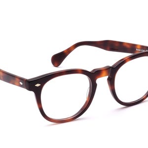 Può includere: Occhiali marroni tartarugati con montatura rotonda. Gli occhiali hanno un design classico con ponte a chiave e dettagli a forma di diamante sulle aste. Le lenti sono trasparenti.