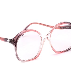 Op de afbeelding: Oversized, roze getinte bril met een helder montuur. De bril heeft een ronde vorm en een subtiel logo op de poot. De monturen gaan over van een donkerdere roze tint naar een lichtere, doorschijnende tint.