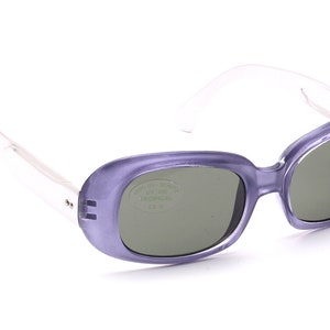 Könnte beinhalten: Lilafarbene Sonnenbrille aus Kunststoff mit einem klaren Gestell und grünen Gläsern. Die Gläser tragen den Aufdruck "UV 400 TROPICAL".
