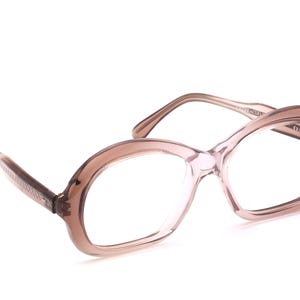 Könnte beinhalten: Eine Vintage-Brille mit einem hellbraunen, ovalen Rahmen. Die Gläser sind klar, und die Bügel haben ein strukturiertes Design. Die Brille ist vor einem weißen Hintergrund platziert.