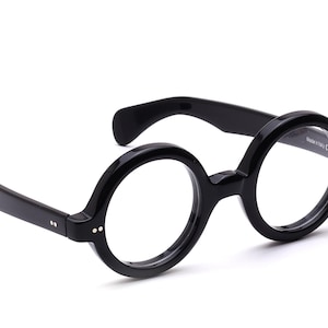 Può includere: Occhiali neri con montatura rotonda e spessa. Il design è classico con un ponte a chiave. Le aste sono dritte e presentano piccoli rivetti decorativi. Sfondo bianco.