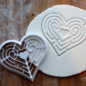 May include: A white heart-shaped cookie cutter with a maze design inside. The cutter is placed on top of a white heart-shaped piece of fondant, also with a maze design.