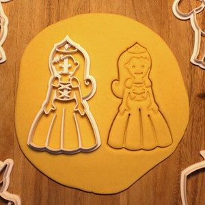 May include: Two white cookie cutters shaped like princesses. The princess on the left has a crown, long hair, and a long dress with a lace-up bodice. The princess on the right has a crown, long hair, and a long dress with a bodice.