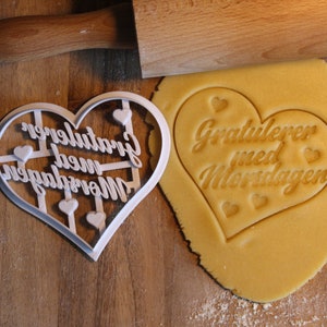 May include: A white plastic heart-shaped cookie cutter with the text "Gratulerer med Morsdagen" in a decorative font. The cookie cutter is on a wooden surface next to a cookie with the same text imprinted on it.