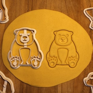 May include: Two white plastic cookie cutters shaped like bears. The bears are sitting upright with their paws in front of them. The cookie cutters are on a yellow surface.