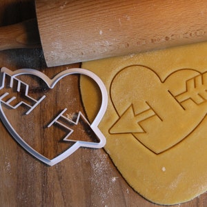 May include: A heart-shaped cookie cutter with an arrow design. The cutter is made of white plastic and is sitting on a wooden surface. A heart-shaped cookie has been cut out of dough using the cutter.