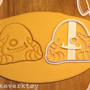 May include: Two white plastic cookie cutters shaped like a cartoon monster with horns and a toothy grin. The cutters are on a yellow surface.