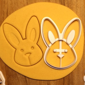 May include: A white plastic cookie cutter in the shape of a bunny rabbit. The cutter is on top of a yellow piece of dough.